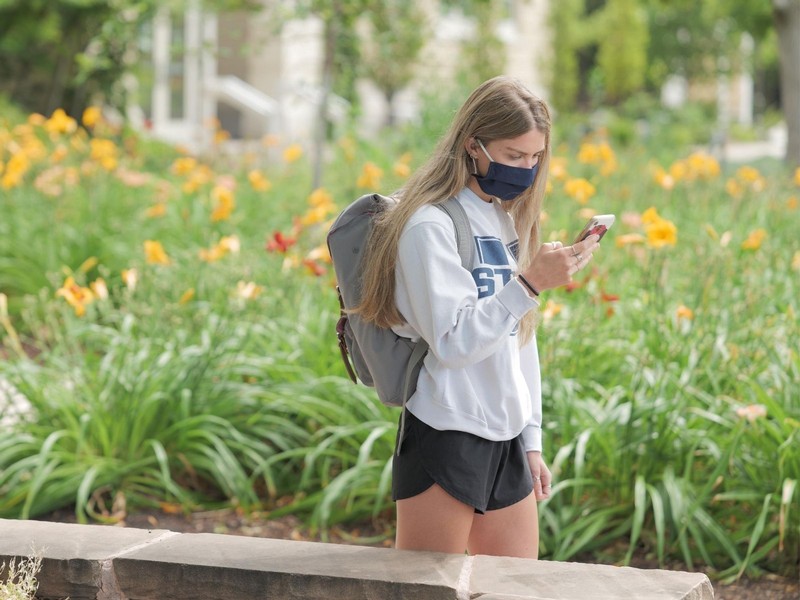A student looks at her phone while walking on campus in her mask.
