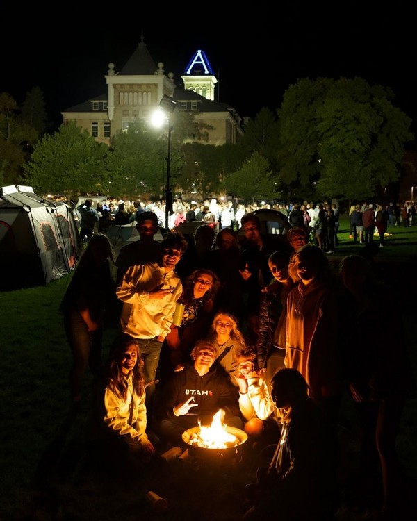 USU students sit in front of a campfire, while camping on the quad at night.