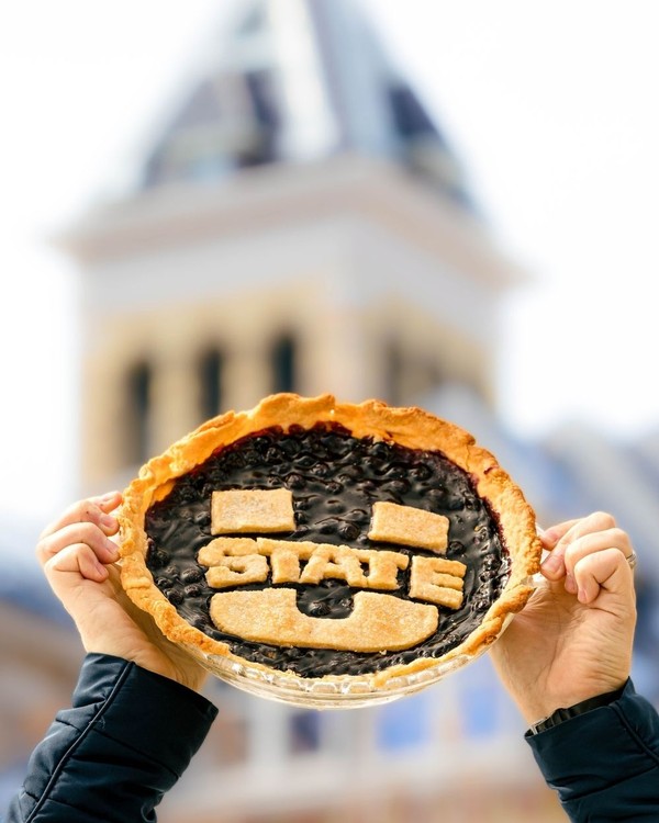 A USU-themed pie is held up in front of Old Main