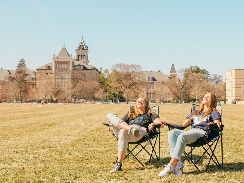 Two female students relax in fold-out chairs on the Quad, soaking in the spring sunshine.