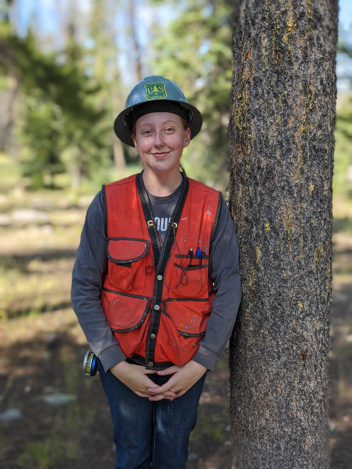 A woman standing in a forest wears a U.S. Forest Service helmet and vest.