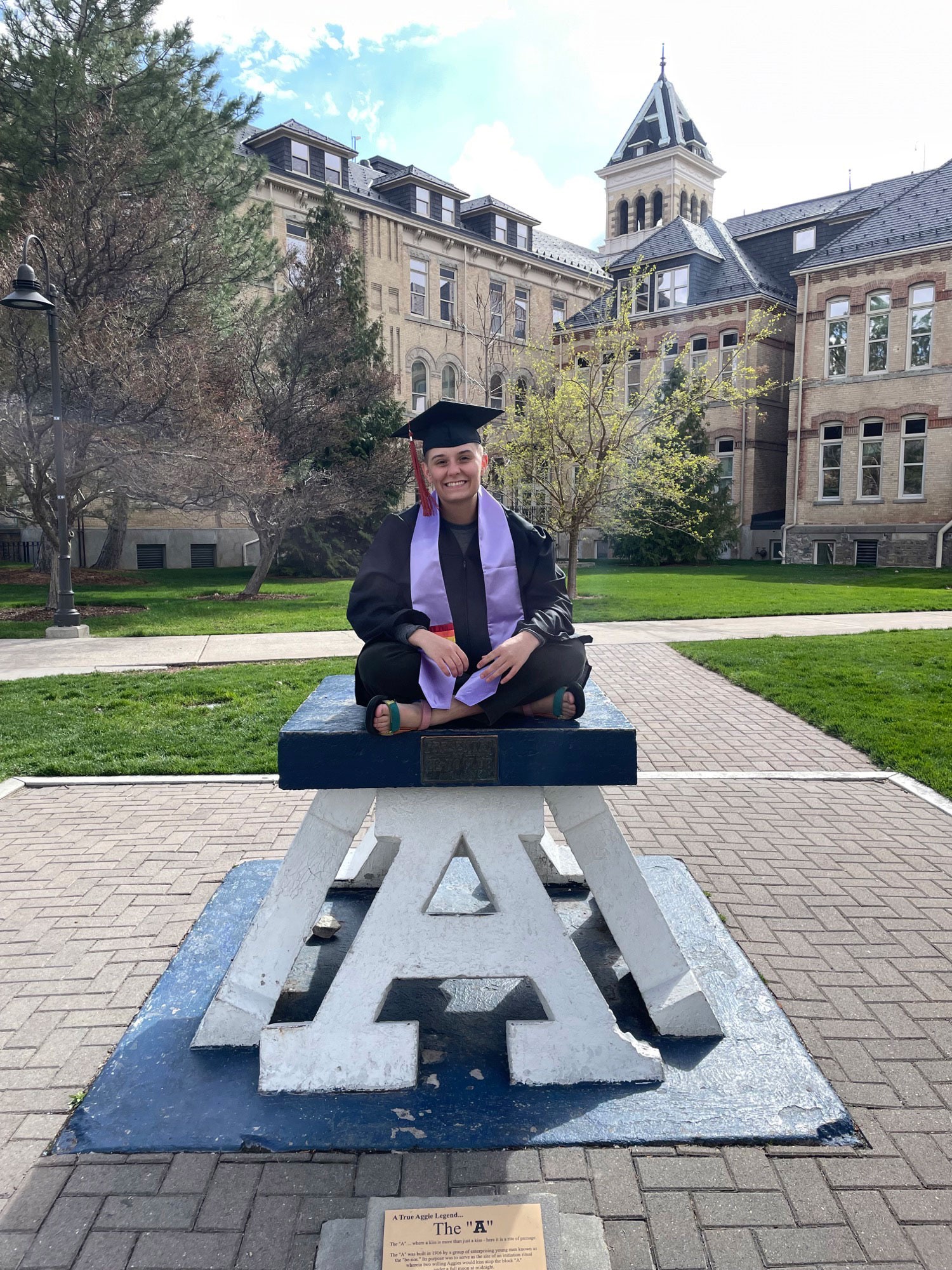 River Johnson sits on the Block A in front of Old Main in graduation robes.